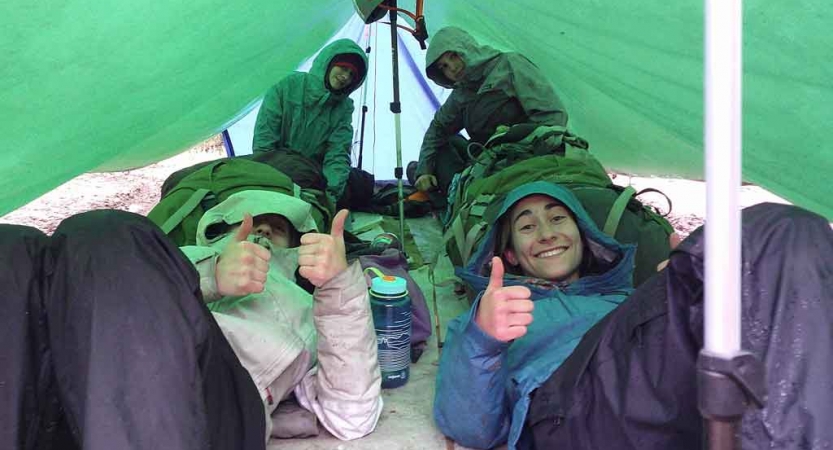 From under a tarp shelter, people smile and give the camera thumbs up.